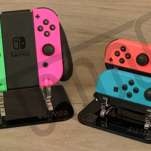 Puede incluir: Tres mandos Joy-Con de Nintendo Switch en soportes de acrílico negro. Los mandos son verde y rosa, rojo y azul.