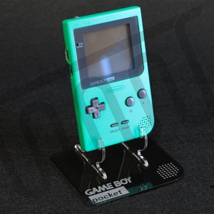 Puede incluir: Una consola de videojuegos portátil Game Boy Pocket de color verde azulado sobre un soporte negro con el texto "GAME BOY pocket" en la parte delantera.