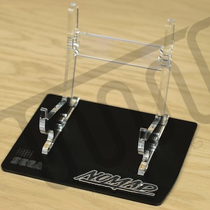 May include: A black acrylic stand with a clear acrylic display for a Sega Nomad handheld games console. The stand has the words "SEGA" and "Nomad" printed on it.