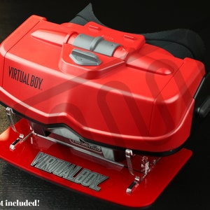以下が含まれることがあります： 透明なアクリル製のスタンドに置かれた赤いバーチャルボーイヘッドセットディスプレイ。スタンドには「VIRTUAL BOY」の文字が印刷されています。本体は含まれていません。