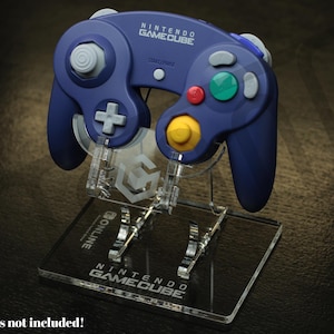 Puede incluir: Un mando azul de Nintendo GameCube expuesto en un soporte de acrílico transparente. El soporte tiene el logotipo y el texto de Nintendo GameCube. El mando tiene botones grises, rojos, verdes y amarillos. El soporte está diseñado para sostener el mando.