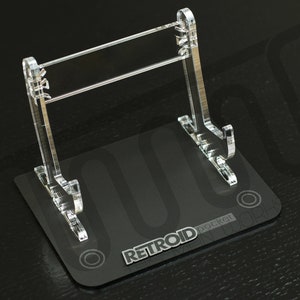 Può includere: Supporto espositivo in acrilico trasparente per un dispositivo di gioco portatile Retroid Pocket. Il supporto ha una base nera con la scritta "RETROID pocket" e una struttura di supporto verticale trasparente. Il supporto è progettato per tenere il dispositivo in posizione verticale.