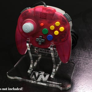 May include: A clear acrylic stand displaying a translucent red Nintendo 64 controller. The controller has gray, blue, green, and yellow buttons. The stand has the Nintendo 64 logo and the text "HORI".