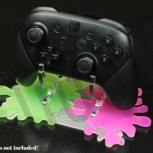 Puede incluir: Un mando de Nintendo Switch negro se muestra en un soporte de acrílico verde y rosa con forma de pintura salpicada. El soporte tiene el texto "Splatoon" y "The Man Works" impreso en él.
