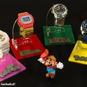 Puede incluir: Cuatro soportes para relojes con bases y logotipos de diferentes colores. Los logotipos son "Pokémon", "Super Mario Bros.", "Zelda" y "Pokémon". Una pequeña figura de Mario está en el centro de la imagen.