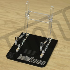 Pode incluir: Um suporte de exibição de acrílico preto com uma tela de acrílico transparente para um cartucho de videogame. O suporte tem o texto "Turbo Express" impresso na base.