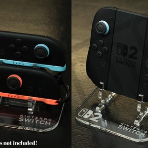 Può includere: Controller Nintendo Switch neri con dettagli blu e arancioni esposti su supporti in acrilico trasparente. I supporti presentano il logo Nintendo Switch e la scritta "Controllers not included!"