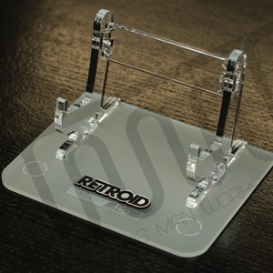 Può includere: Supporto espositivo in acrilico trasparente per un dispositivo di gioco portatile. Il supporto ha una base rettangolare con il testo "RETROID pocket / Flip" e un logo. Design chiaro e minimalista.