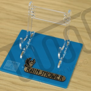 Puede incluir: Un expositor de acrílico transparente con una base azul. La base tiene el texto "GAME & WATCH" y "RAIN SHOWER" impreso en ella.