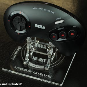 May include: Black Sega Mega Drive controller on a clear acrylic stand. The controller has a directional pad, start button, and A, B, and C buttons. The stand is labeled "SEGA 16-BIT MEGA DRIVE" and "ONLINE".