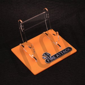 Puede incluir: Un expositor de acrílico transparente con dos brazos para sostener objetos. El expositor tiene una base naranja con las palabras "FIRE ATTACK" impresas en él.