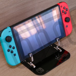 Puede incluir: Un soporte negro con una consola Nintendo Switch en la parte superior. La consola está en modo de sobremesa con un mando Joy-Con azul y rojo conectado. El soporte tiene las palabras "Nintendo Switch" impresas.