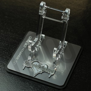 May include: A clear acrylic stand for a Game Boy Advance SP, with a silver Kingdom Hearts logo on the base.
