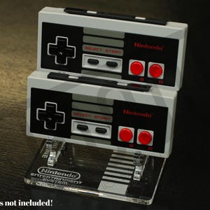 May include: Stacked display stands for Nintendo Entertainment System controllers. The stands are clear acrylic with a black and grey base. The controllers are white, black, and grey with red buttons and the Nintendo logo.