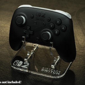 以下が含まれることがあります： 黒色のNintendo Switch Proコントローラーが透明なアクリルスタンドに置かれています。スタンドにはNintendo Switchのロゴとテキストが表示されています。コントローラーには白いアクセントとボタンのラベルが付いています。スタンドはコントローラーを立てて保持するように設計されています。