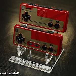 Könnte beinhalten: Zwei rote und goldfarbene Gamecontroller im Retro-Stil, gestapelt auf einem transparenten Acrylständer. Die Controller haben schwarze Knöpfe und ein kreuzförmiges Steuerkreuz. Der Ständer trägt den Text "FAMILY COMPUTER Nintendo".