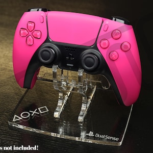 Op de afbeelding: Een felroze DualSense-controller staat op een heldere acryl standaard. De controller heeft zwarte accenten en roze knoppen. De standaard heeft de PlayStation-symbolen en de woorden "DualSense" erop gedrukt. De tekst "Controllers not included!" staat onderaan.