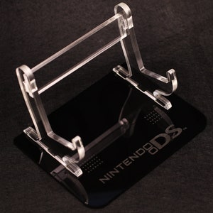 Op de afbeelding: Transparante acryl Nintendo DS gameconsole standaard. De standaard heeft een rechthoekige basis met het Nintendo DS-logo en een transparante, schuine ondersteuningsstructuur. Ontworpen om een Nintendo DS gameconsole te ondersteunen.