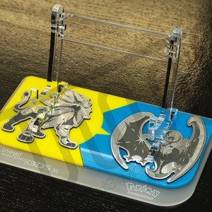 Puede incluir: Un soporte de acrílico transparente con una base amarilla y azul que presenta personajes de Pokémon. El soporte está diseñado para sostener una consola Nintendo 3DS XL. El texto "new Nintendo 3DS XL" está impreso en la base.