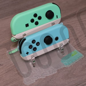 Puede incluir: Dos mandos Nintendo Switch Joy-Con de color azul turquesa y blanco en un soporte de plástico transparente. Los mandos se muestran sobre una superficie de madera.