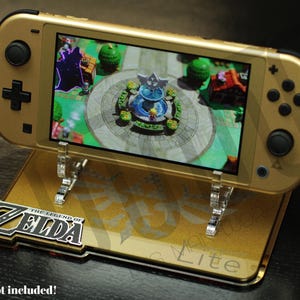 Può includere: Console Nintendo Switch Lite dorata su un supporto abbinato, che mostra una scena di gioco. Il supporto presenta il logo "The Legend of Zelda". La console è supportata da supporti in acrilico trasparente. Il testo "Console non inclusa!" è visibile.