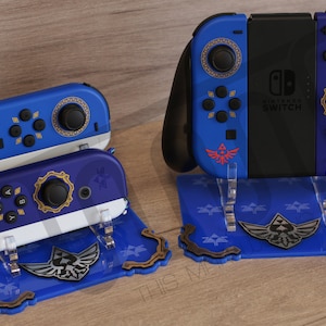 May include: A blue and gold Nintendo Switch console and two blue and gold Joy-Con controllers with a Triforce design on a blue stand. The stand has a Triforce design and the text "Nintendo Switch" on the console.