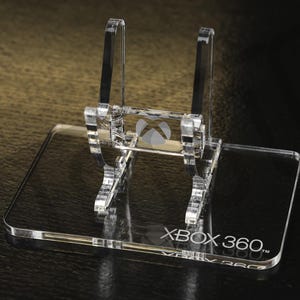 May include: Clear acrylic Xbox 360 console stand. The stand features the Xbox logo and the text "XBOX 360." The stand is designed to hold the console upright. The stand is made of clear acrylic.