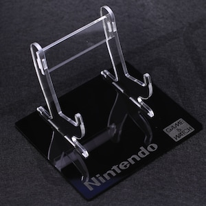 Puede incluir: Un soporte de acrílico transparente para una consola portátil Nintendo Game & Watch. El soporte tiene una base negra con las palabras "Nintendo" y "GAME & WATCH" grabadas en ella.