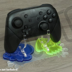 Puede incluir: Un mando Nintendo Switch Pro negro en un soporte de acrílico transparente con un diseño azul y verde. El soporte presenta un diseño de calamar estilizado. El texto "¡Mandos no incluidos!" es visible en la parte inferior de la imagen.