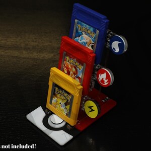 May include: A red, white, and black game cartridge display stand holding three colourful Pokémon game cartridges. The stand features circular icons and the text "Cartridges not included!"