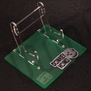 May include: A clear acrylic stand with a green base for displaying Game & Watch: Donkey Kong. The base features the Game & Watch logo and a silver Donkey Kong logo.