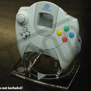 May include: A light grey Dreamcast controller on a clear acrylic stand. The controller has coloured buttons and a directional pad. The stand has the Dreamcast logo and the text "Controllers not included!"