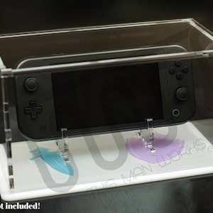 Puede incluir: Una vitrina transparente de acrílico con una consola Nintendo Switch negra en su interior. La vitrina tiene una base blanca con un diseño azul y morado. El texto "THIS MAN WORKS" es visible en la base. ¡Consola no incluida!