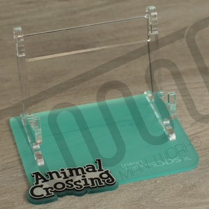 Può includere: Un supporto in acrilico trasparente con una base turchese per una Nintendo 3DS XL. La base ha la scritta "Animal Crossing" stampata.