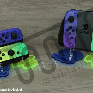 Puede incluir: Dos mandos Joy-Con de Nintendo Switch con diseños personalizados en morado y verde, expuestos en soportes de acrílico transparente con forma de pulpo. Los soportes están sobre una superficie de madera. "This Man Works" está impreso en la superficie. "¡Mandos no incluidos!" está escrito en una señal de advertencia en la parte inferior de la imagen.