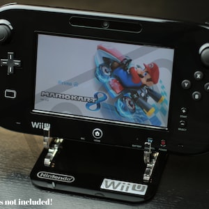 Puede incluir: Una consola Nintendo Wii U negra con un soporte. La consola está mostrando el juego Mario Kart 8 en la pantalla. La consola tiene un diseño negro y plateado con un logotipo de Nintendo en la parte delantera.
