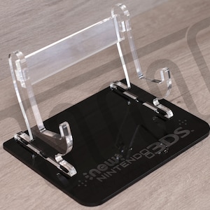 Puede incluir: Un soporte de acrílico transparente para una consola de juegos portátil Nintendo 3DS. El soporte tiene una base negra con el texto "new Nintendo 3DS" impreso en él.