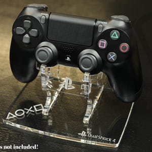 Puede incluir: Un mando DualShock 4 negro sobre un soporte de acrílico transparente. El soporte muestra el logotipo de PlayStation y el texto "DualShock 4". El mando tiene la disposición estándar de botones, sobre una superficie oscura.