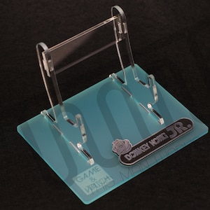 May include: A clear acrylic video game display stand with a blue frosted base. The base has the text "GAME & WATCH" and "DONKEY KONG JR." printed on it.
