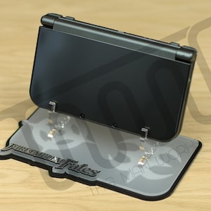 May include: A black Nintendo 3DS handheld gaming console on a clear acrylic stand with a gray base. The base has the words "Fire Emblem Fates" printed on it.