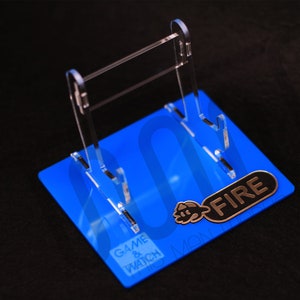 Puede incluir: Un soporte de acrílico transparente para una consola de juegos portátil Game & Watch Fireman. El soporte está sobre una base azul con las palabras "Game & Watch" y "Fireman" impresas en él. El soporte tiene una etiqueta de metal dorado con la palabra "FIRE" impresa en ella.