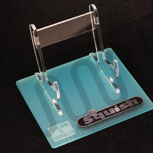 Puede incluir: Un soporte de acrílico transparente con tres ranuras para exhibir consolas Game & Watch. El soporte tiene una base azul esmerilada con el texto "Game & Watch" y el logotipo "Squish This Gaming" impreso en él.