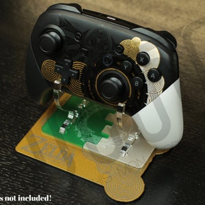 Peut inclure: Un support de manette Nintendo Switch de couleur or et noir avec un design Zelda. Le support a une base verte et or avec un motif circulaire. Le texte "ZELDA" est imprimé sur la base. Le texte "A Controllers not included!" est imprimé sous le support.