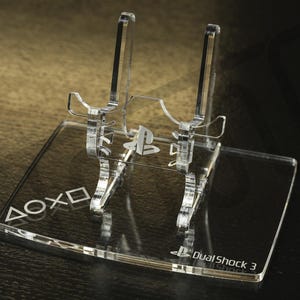 Puede incluir: Soporte transparente de acrílico para mando DualShock 3. El soporte presenta el logotipo de PlayStation y las palabras "ΔΟΧΟ" y "DualShock 3" grabadas en la base. El soporte está diseñado para sujetar un mando en posición vertical.