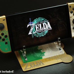 Op de afbeelding: Een gouden Nintendo Switch-console met een groen-gouden Zelda: Tears of the Kingdom-ontwerp op de linker Joy-Con en een wit-gouden ontwerp op de rechter Joy-Con. De console staat op een heldere acrylstandaard met een goud-groen Zelda: Tears of the Kingdom-ontwerp.