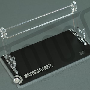 Peut inclure: Un support en acrylique transparent pour un appareil de jeu portable Steam Deck. Le support a une base noire avec le texte "STEAM DECK" et "THIS MAN WORKS" imprimé dessus.