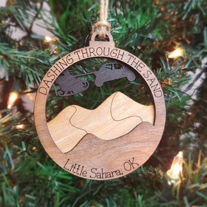 May include: A wooden Christmas ornament with a brown background and a light brown cutout of a dune landscape. The ornament reads "Dashing Through The Sand Little Sahara, OK".