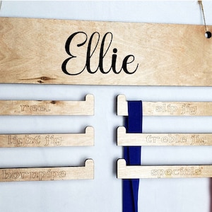 May include: A wooden medal display rack with the name "Ellie" engraved at the top. The rack has five slots for hanging ribbons, each labeled with a different type of fishing lure: "reel", "light jig", "worm pipe", "slip jig", "treble jig", and "specials".