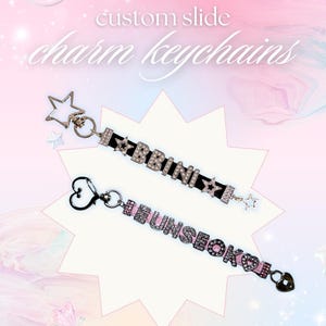 Pode incluir: Dois chaveiros de charme deslizantes personalizados. Um preto com letras prateadas que soletram "BBBINI" e um pingente de estrela. O outro é rosa com letras prateadas que soletram "HEUNSEOK" e um pingente de coração. O fundo é rosa e azul.