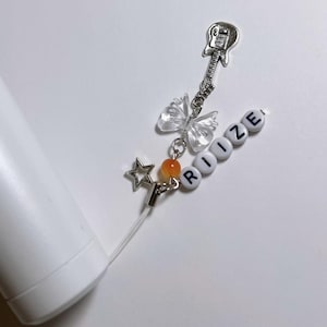 May include: A white phone charm with a silver guitar charm, a clear butterfly charm, a star charm, and orange bead. The charm also includes white letter beads spelling out the word "RIIZE".